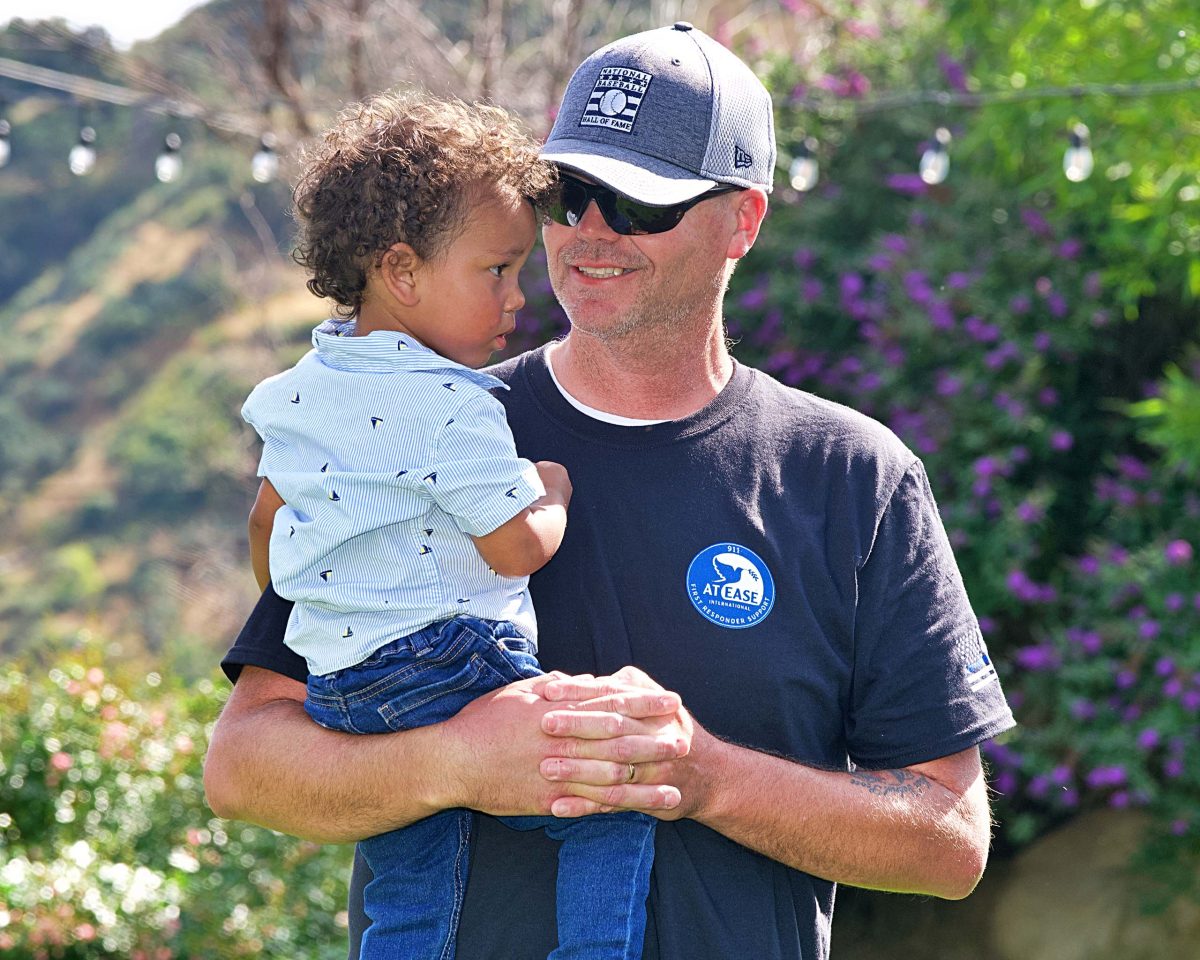 A first responder wearing a 911 At Ease International tshirt while holding his grandson